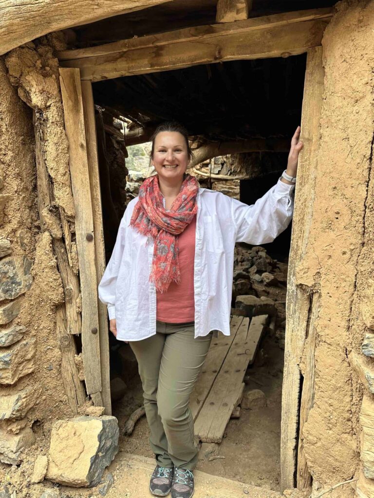 Claire wearing a shirt over hiking gear stood in the doorway for a mud building in an abandoned village. The building has seen better days