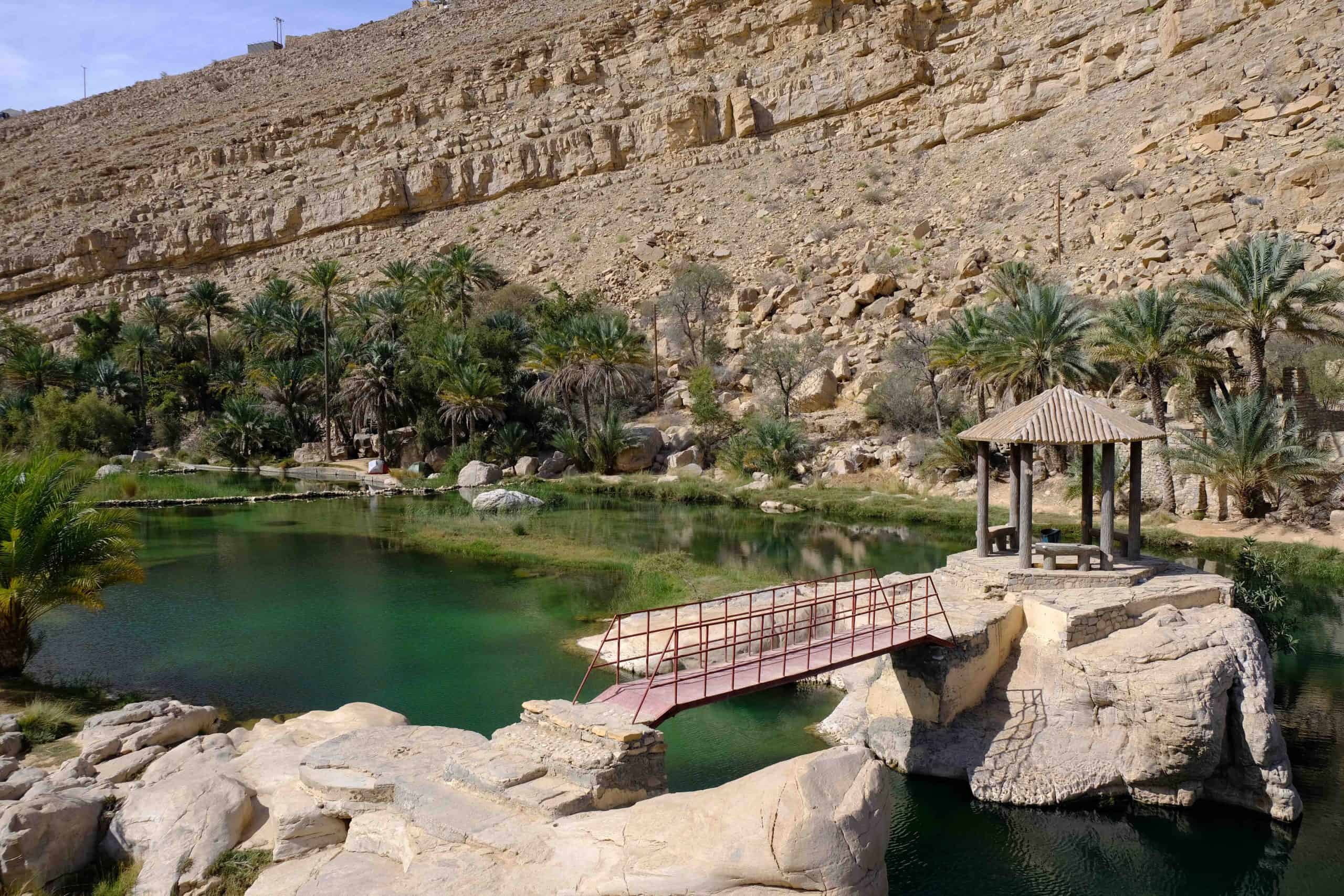 View of the first pool at Wadi Bani Khalid with a bridge and canopy in the foreground. The wadi water looks clear and inviting. The sand coloured valley wall rises in the background