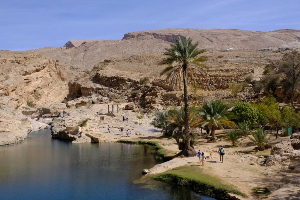 View into Wadi Bani Khalid where the family are walking to one side of the large open pool near the entrance. There are palm trees and people in the distance
