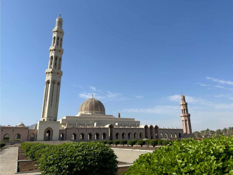 The front of Sultan Qaboos Grand Mosque where there are green bushes. You can see the tallest minaret, dome and another of the four smaller minarets against a blue sky