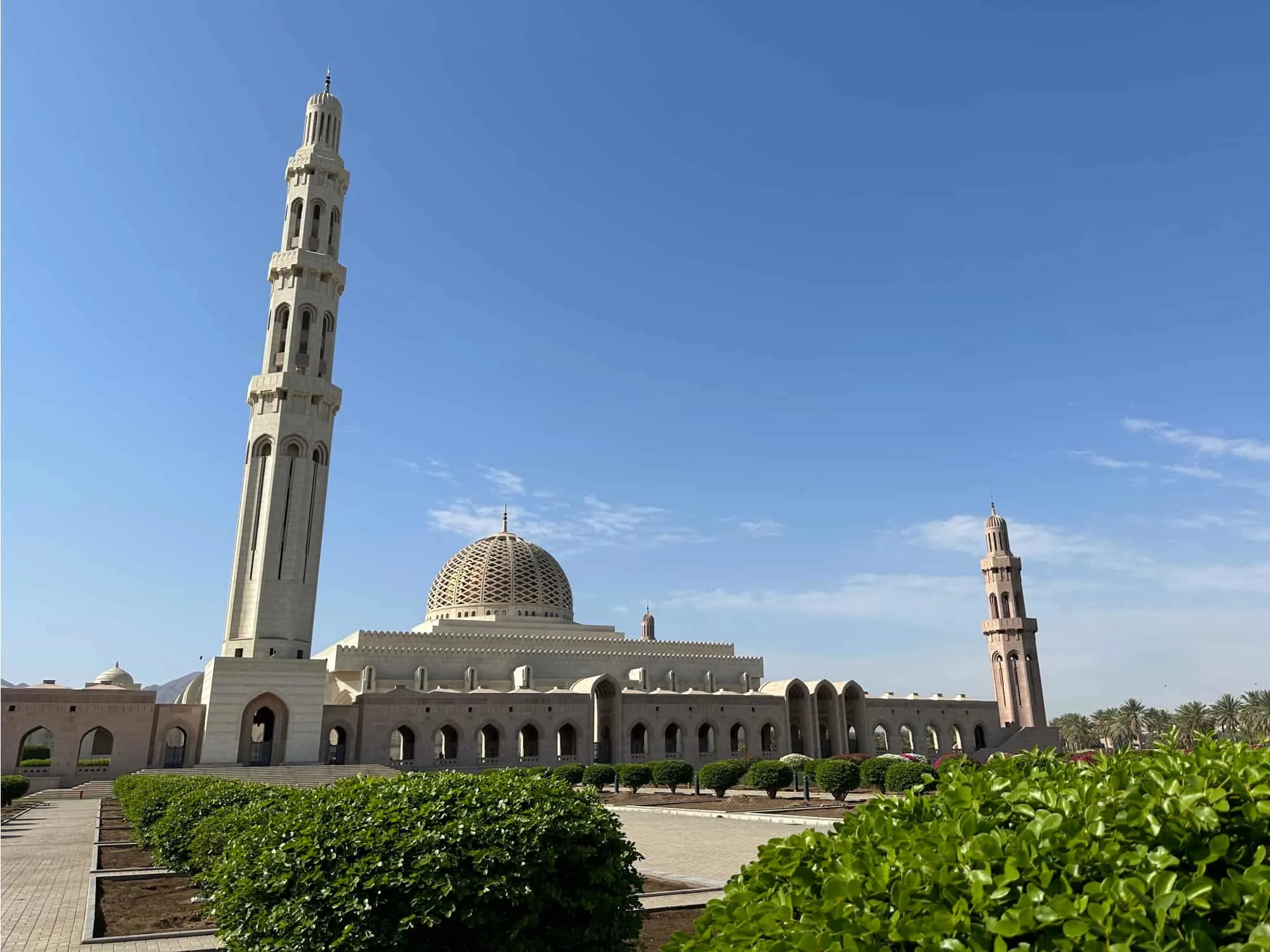 The front of Sultan Qaboos Grand Mosque where there are green bushes. You can see the tallest minaret, dome and another of the four smaller minarets against a blue sky