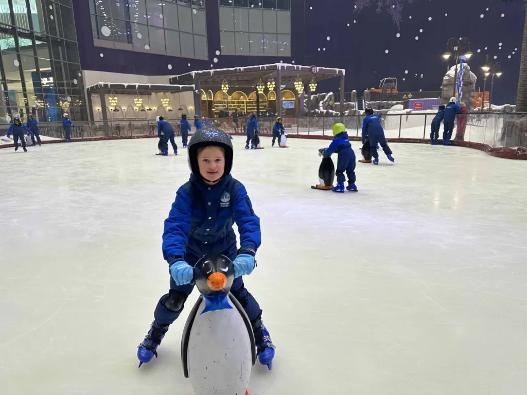 Child in snow suit ice skating with penguin skating aid