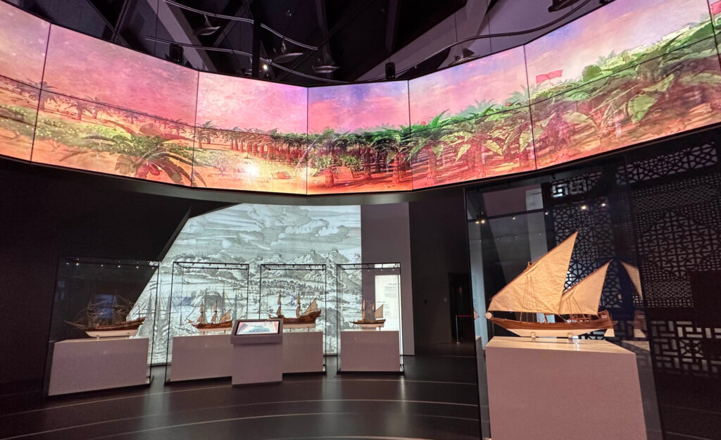 A circular display screen hangs in the air above displays about Oman's maritime history