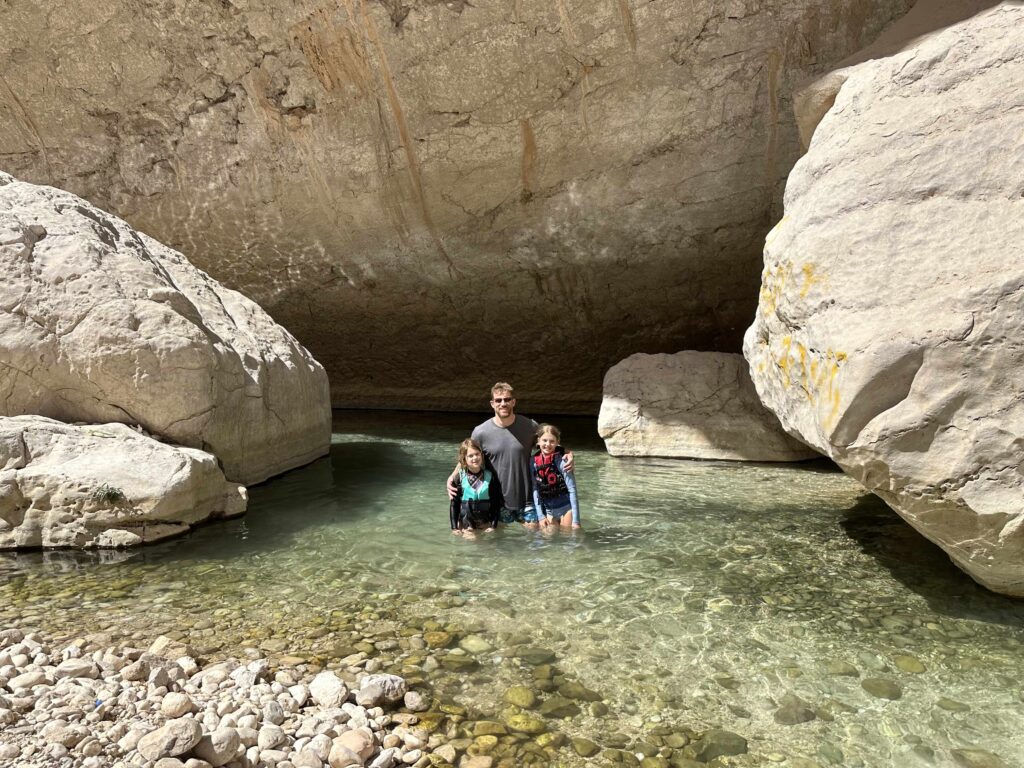 My husband and out children stood in water at Wadi Bani Khalid. They are wearing swimming clothes and life jackets