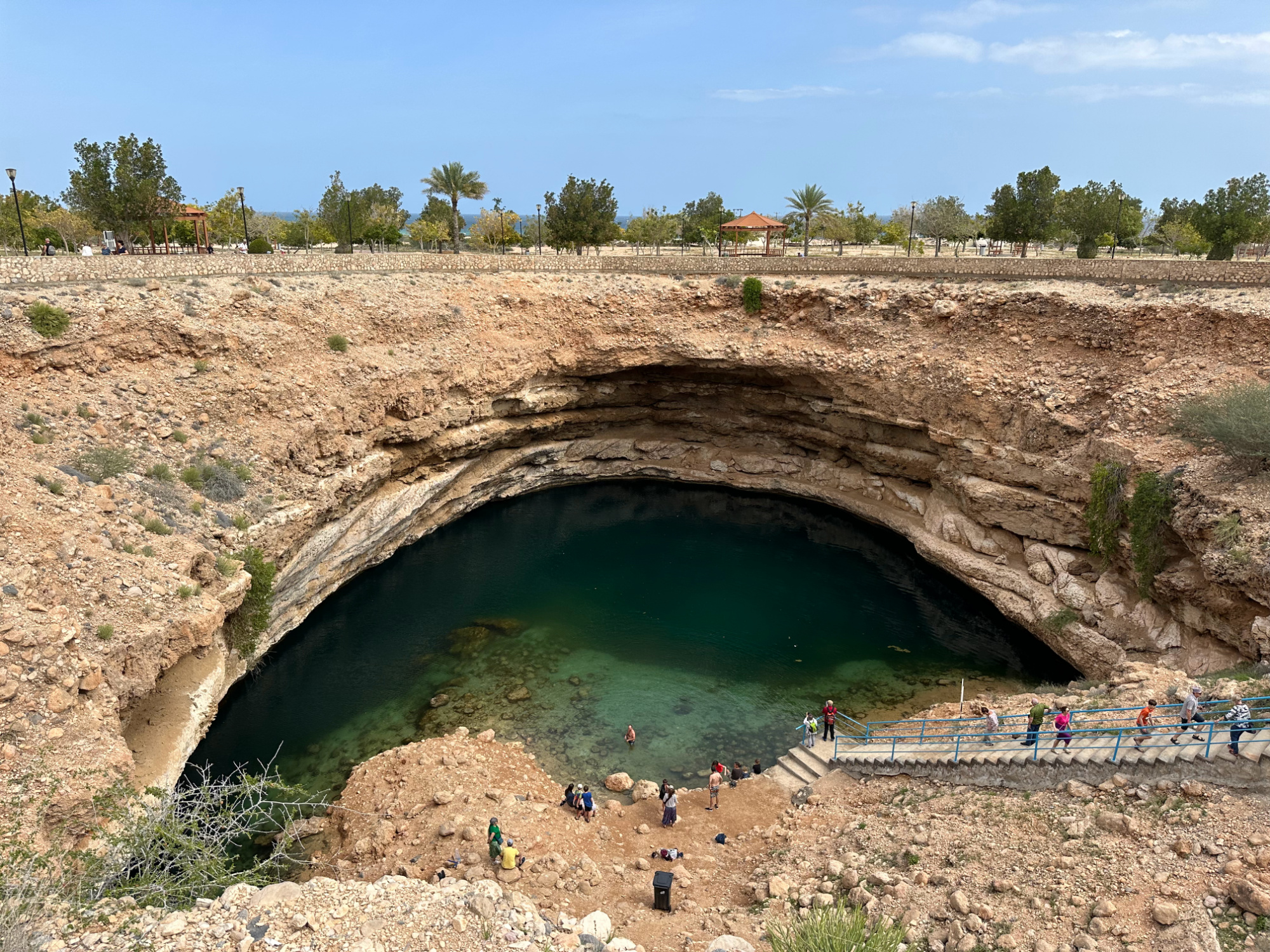 A view of Bimmah Sinkhole from the rim. The sand colour cliffs rise on either side and people walk down the concrete steps towards the water