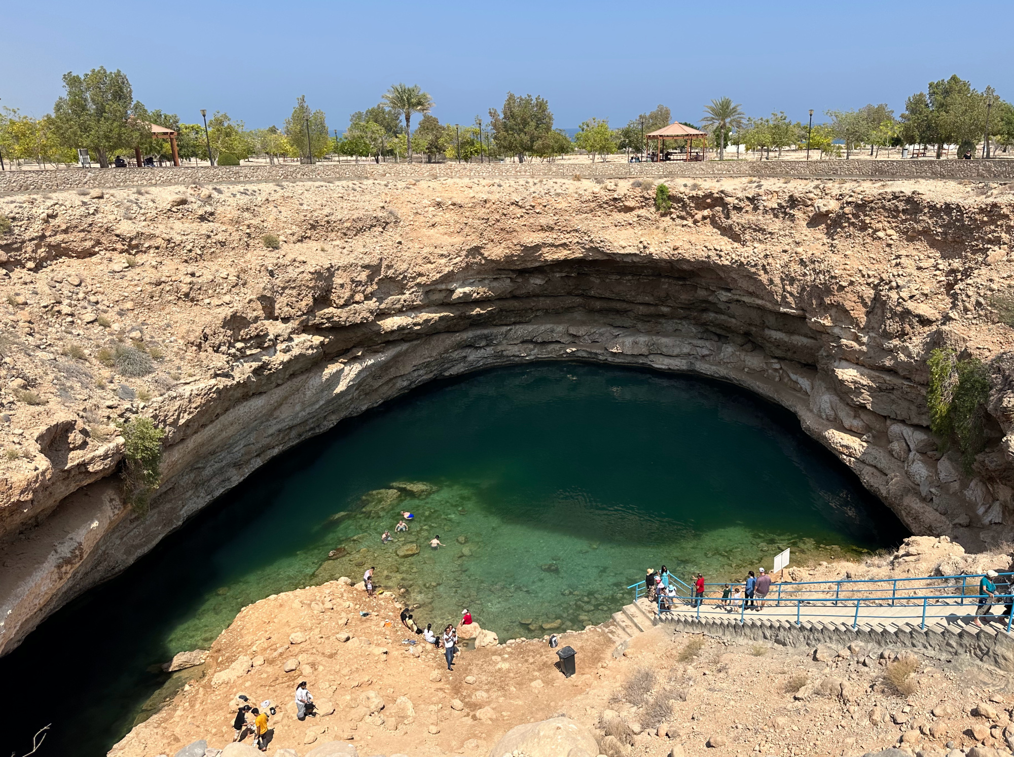 A view of Bimmah Sinkhole from the rim. The sand colour cliffs rise on either side and people walk down the concrete steps towards the water. It's possible to see the water reaching around the side of he mud back to where there is a cave