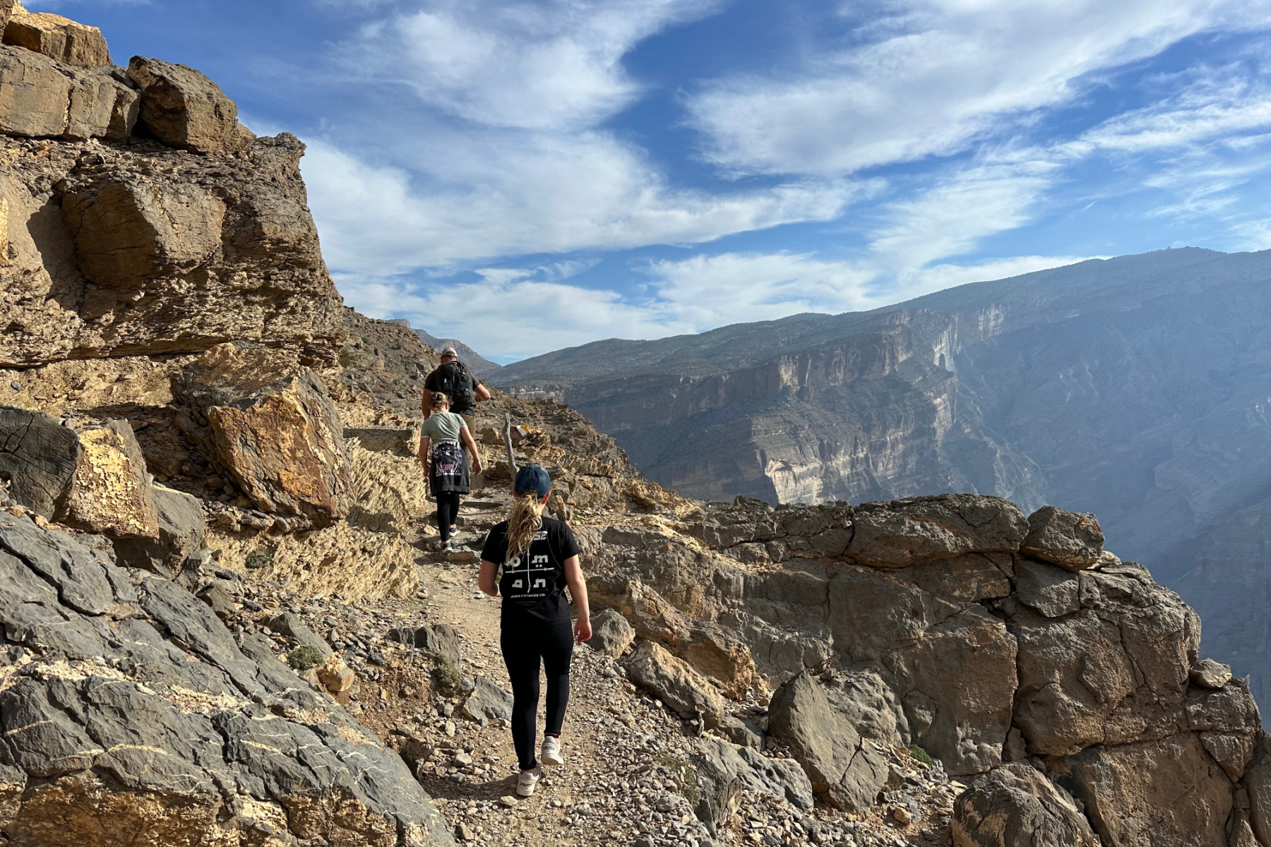 My family walking the path along Oman's Grand Canyon. There is a cliff to their left and a canyon drop to their right. The path is narrow and rocky