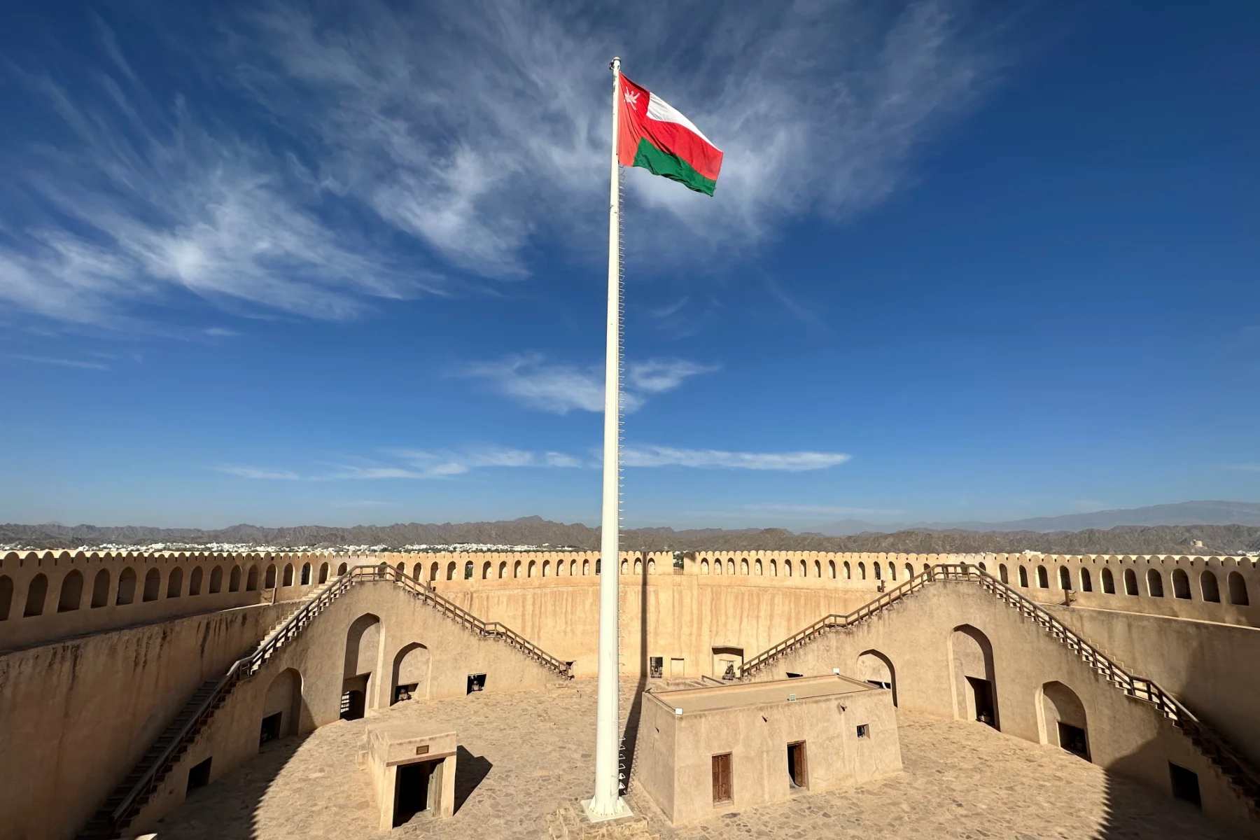 A flag pole flying the Omani flag at the centre of the fort. Beyond you can see the Hajar Mountains