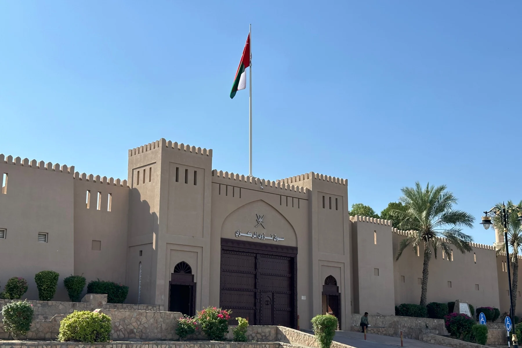 The outside of Nizwa souq. There are high sand coloured walls and dark wood gates. The Omani flag flies above them