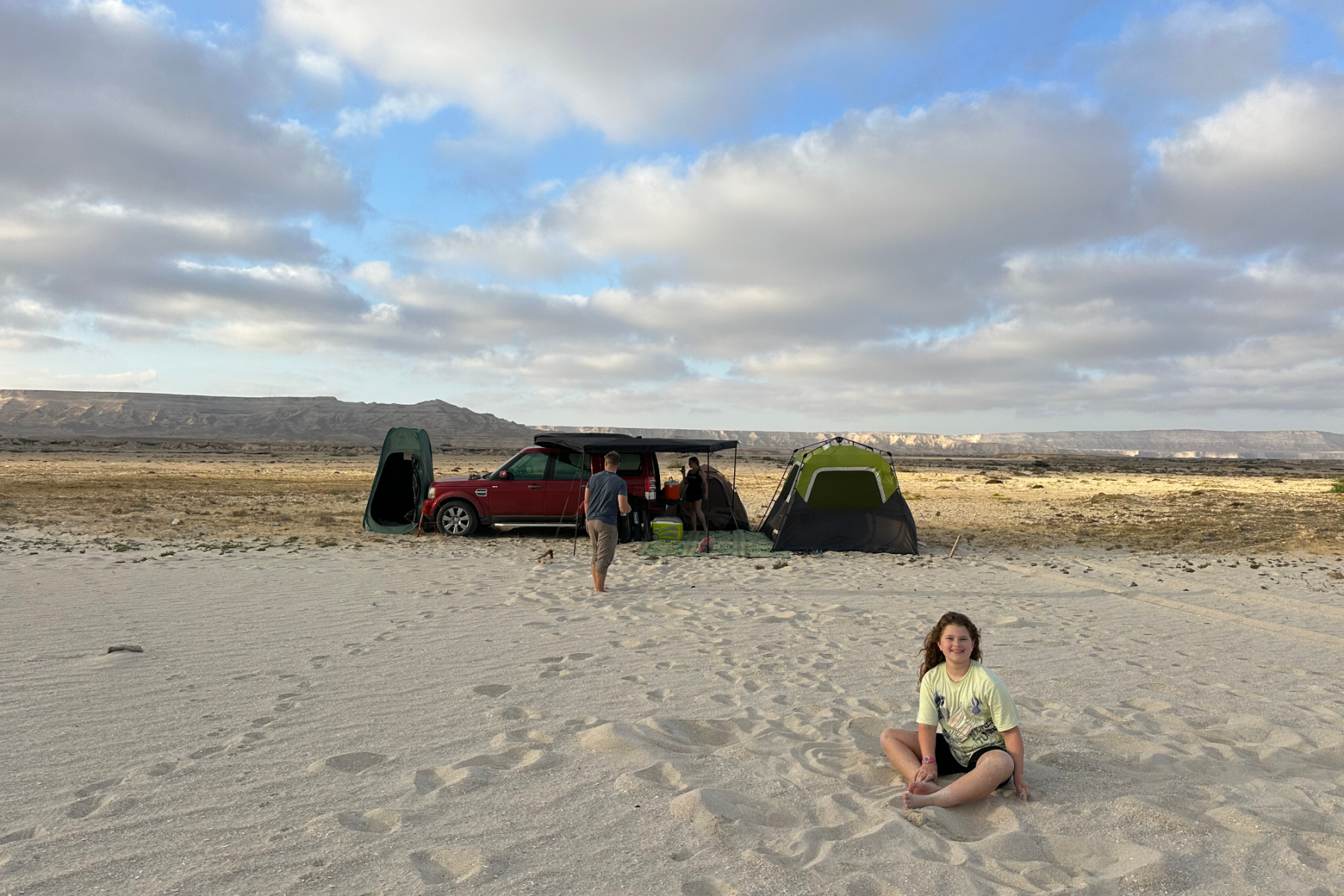One of our daughters set on a sandy beach in front of our Land Rover and tents. In the distance there are mountains