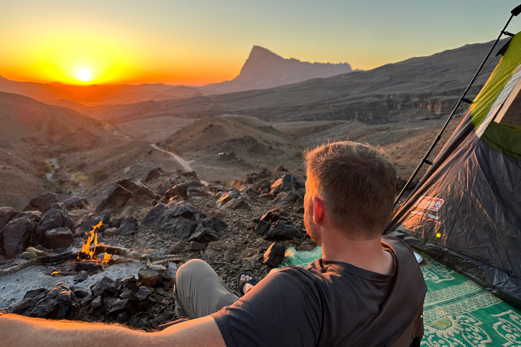 My husband sat on a camping chair beside our tent. In front of him the sun is setting to the left of the distinctive outline of Jebel Misht