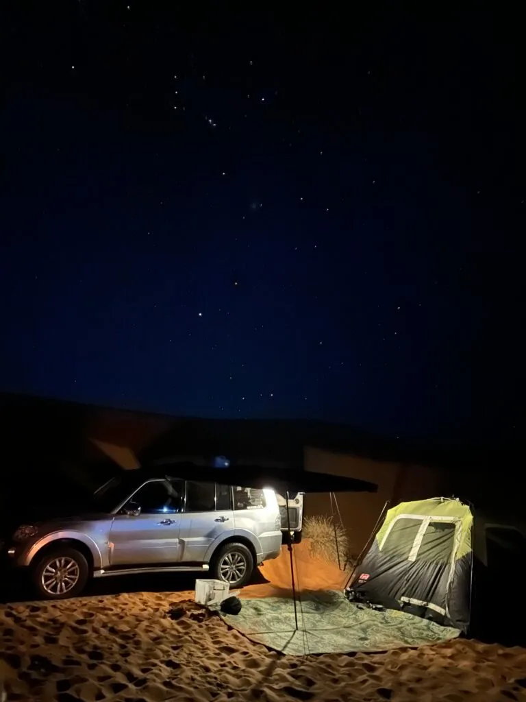 Our car and tent illuminated in the desert at night. Above there are stars in the dark blue sky
