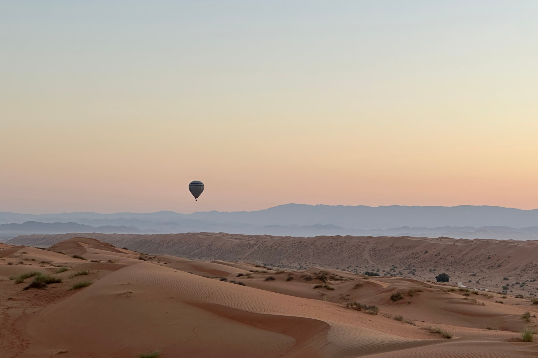 A hot air ballon hanging in the sky over Wahiba Sands desert at dawn. The Hajar Mountains are in the distance and the sand dune are below