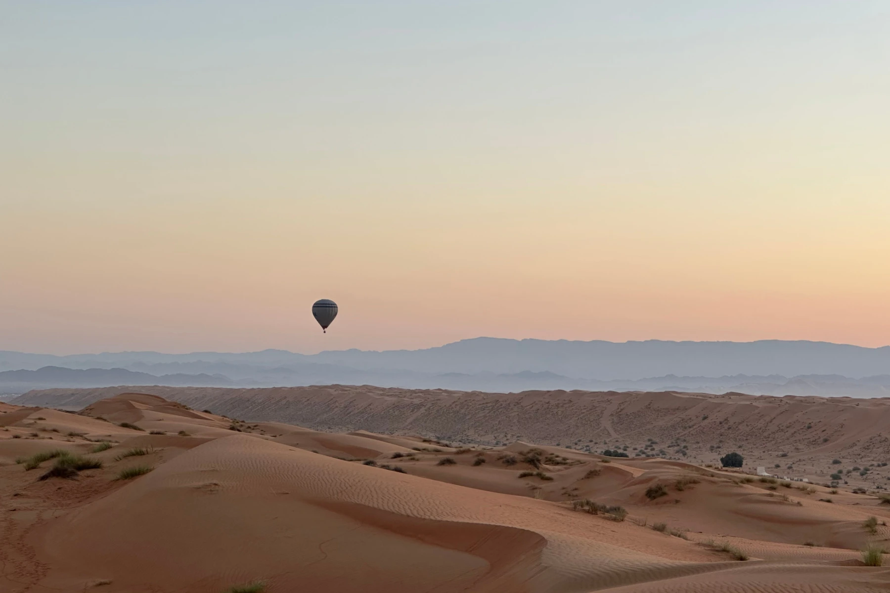 A hot air ballon hanging in the sky over Wahiba Sands desert at dawn. The Hajar Mountains are in the distance and the sand dune are below