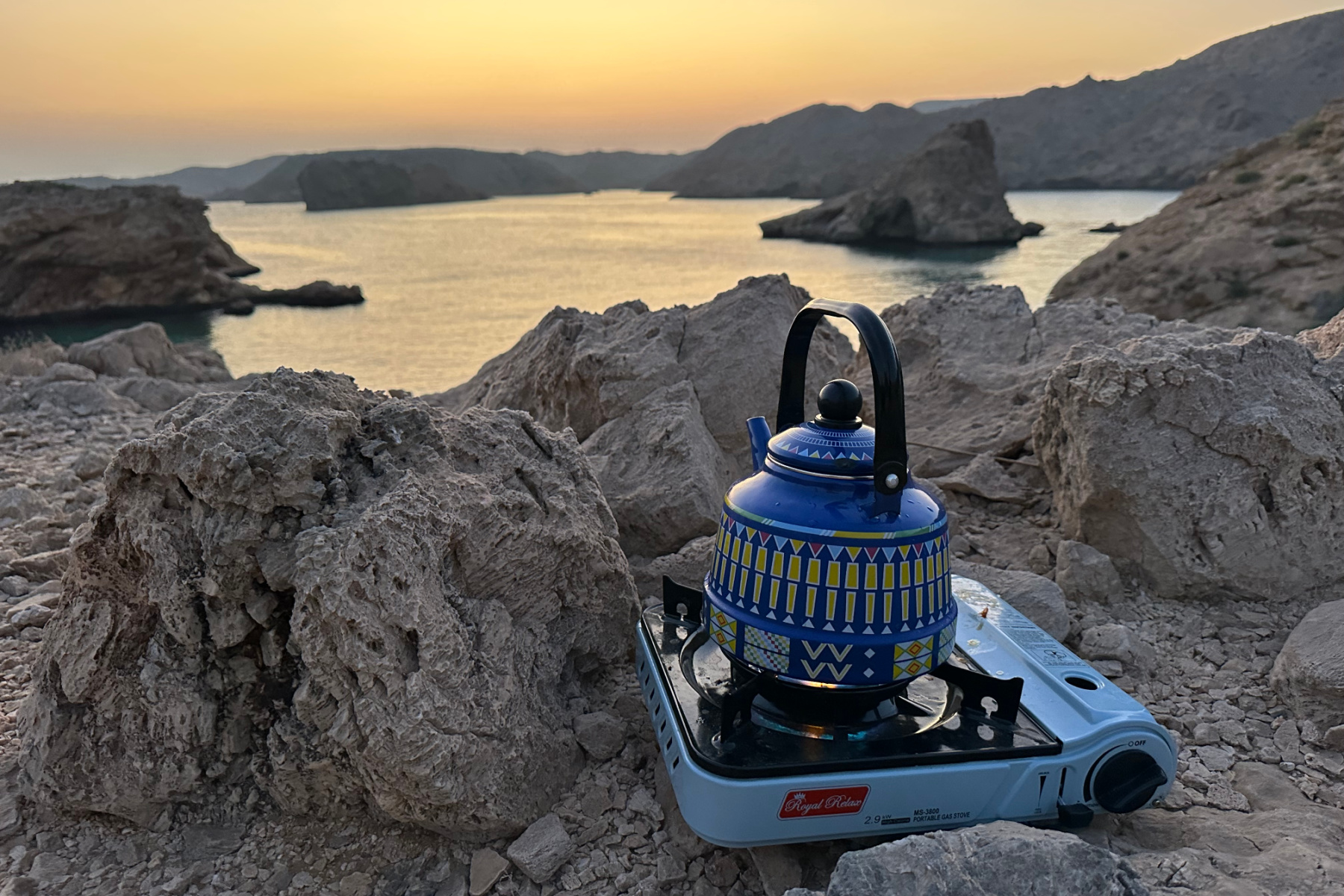 A patterned tin kettle is bowling on a camping stove in front of a view of the Bander Khayran coves to the south east of Muscat. It is dawn and everything is in an orange light