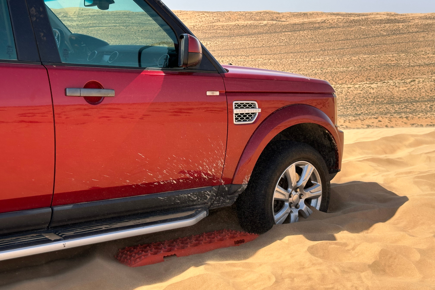 Orange sand board behind the front wheel of a red Landrover Discovery in the desert