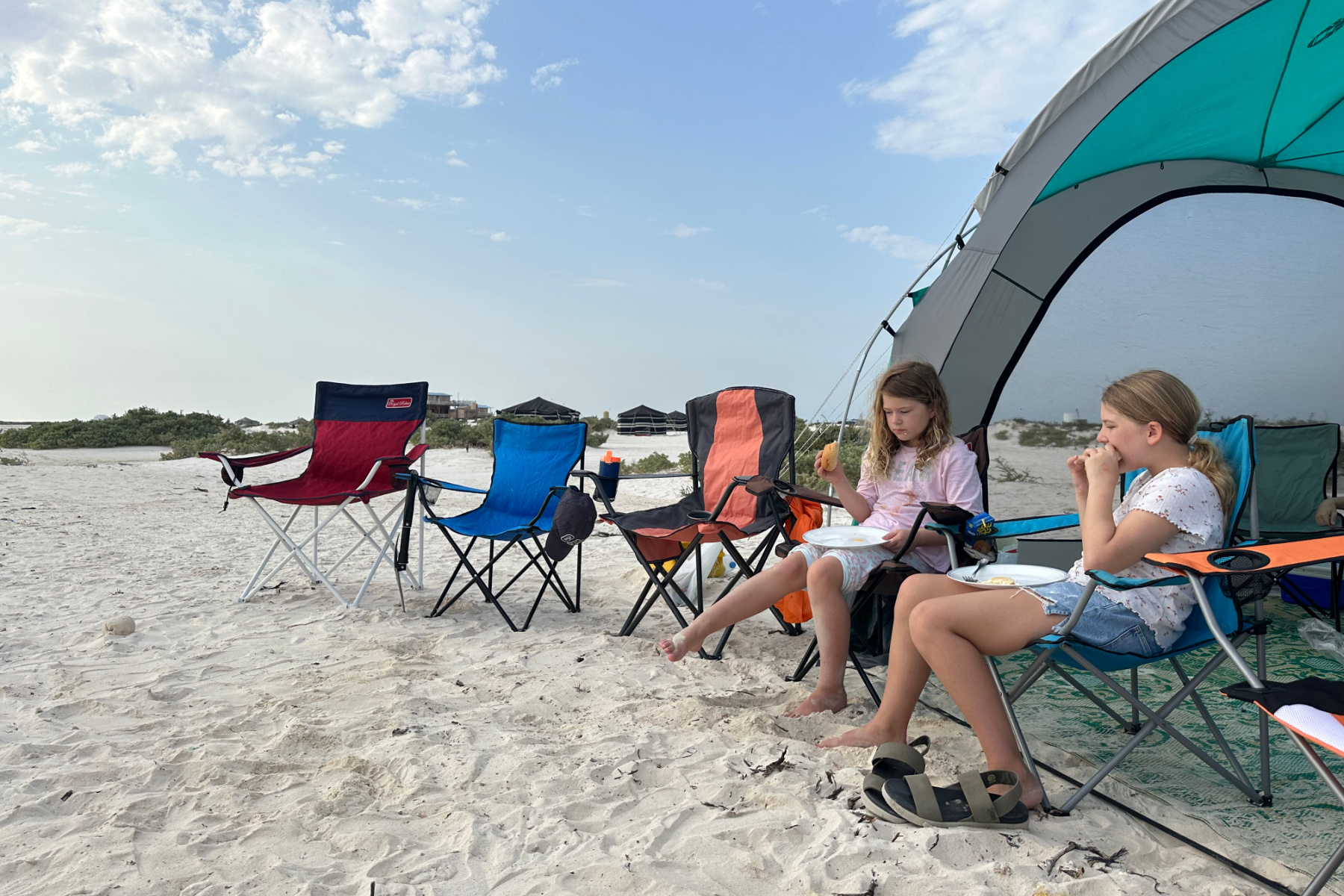 My daughters eating sandwiches while sat on camping chairs in front of a gazebo on the white sands of Barr Al Hikman