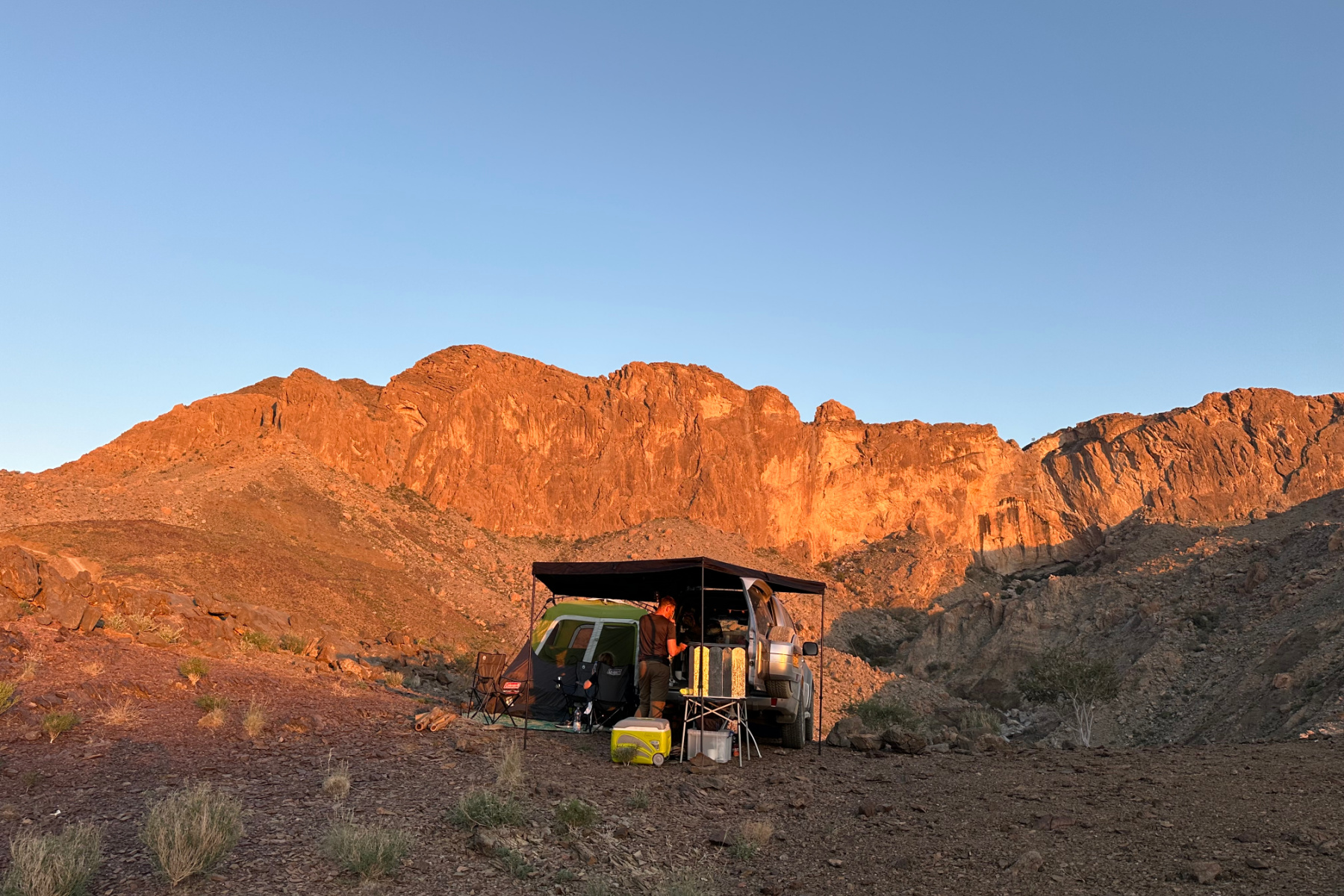 Our car and tent in front of a view of the Hajar Mountains at Wadi Damm. The cliffs are red as the sun is setting behind the camera