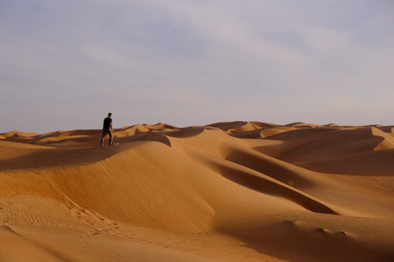 My husband walking across the sand dune in Wahiba Sands at sun rise. The sky is slightly pink
