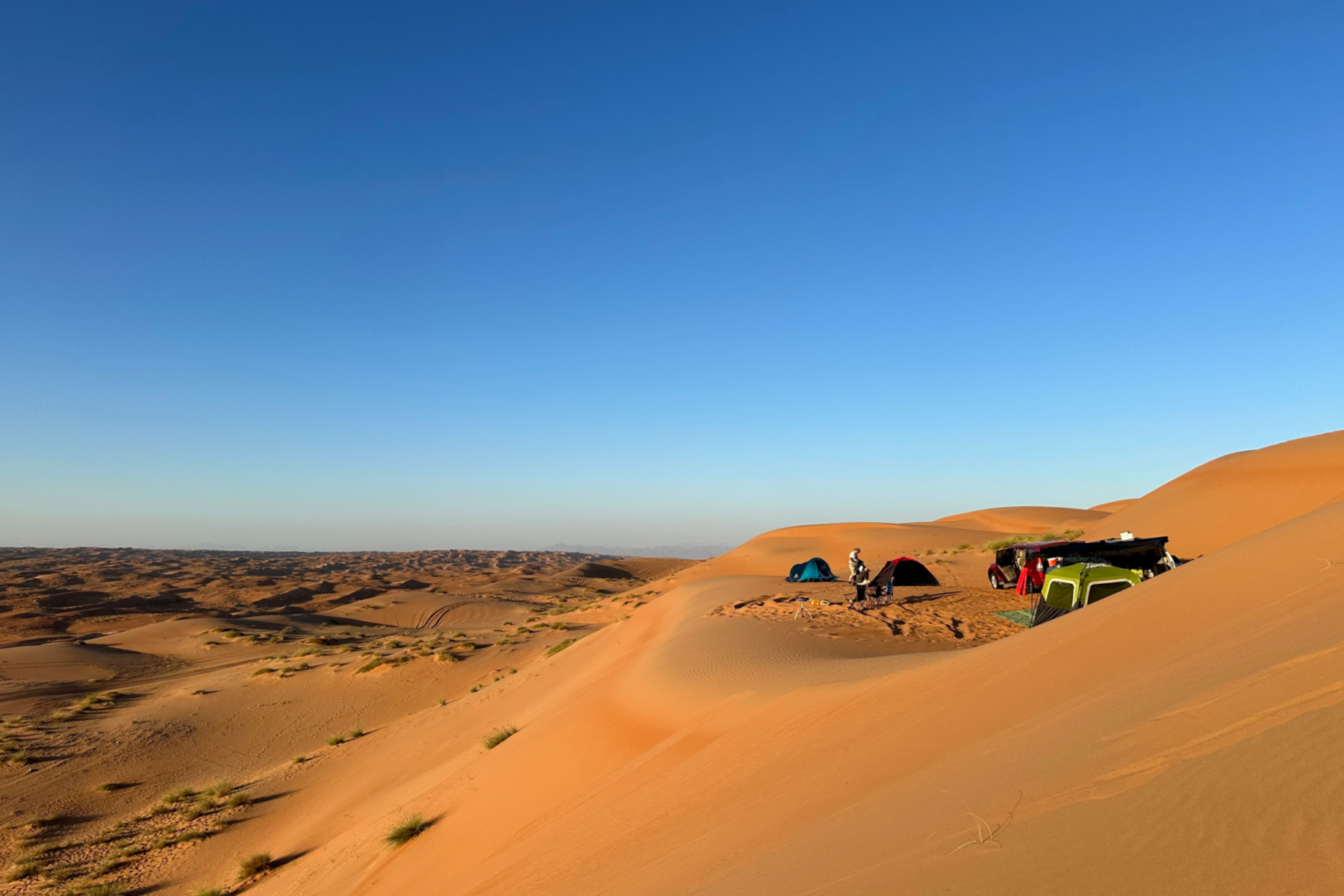 A group of tents pitched in a balcony-like space at the top of a sand dune in Wahiba Sands. It's possible to see all the way down the valley to the Hajar Mountains