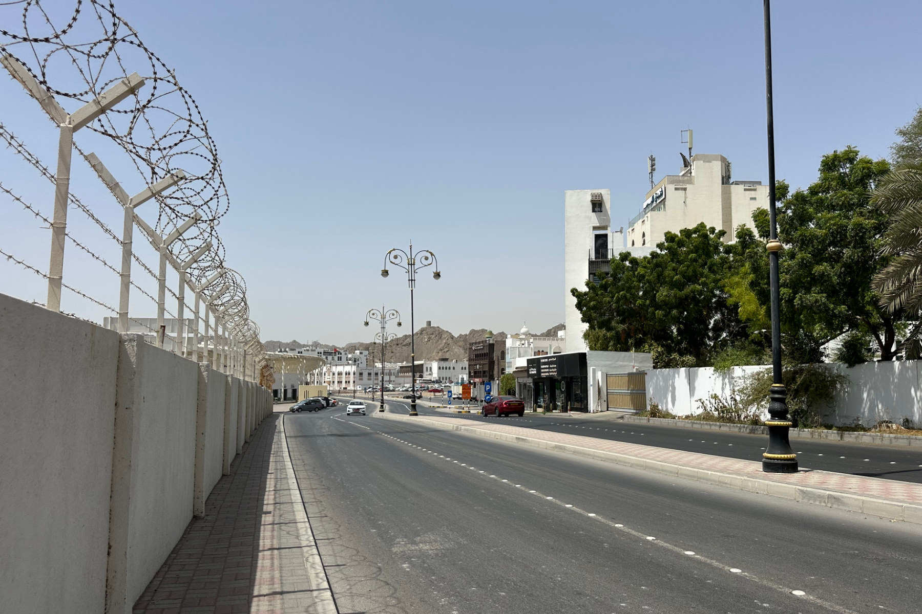 The road from Sultan Qaboos Port towards Muttrah corniche. There is a barbed wire fence on the left and a wall with trees behind it on the right