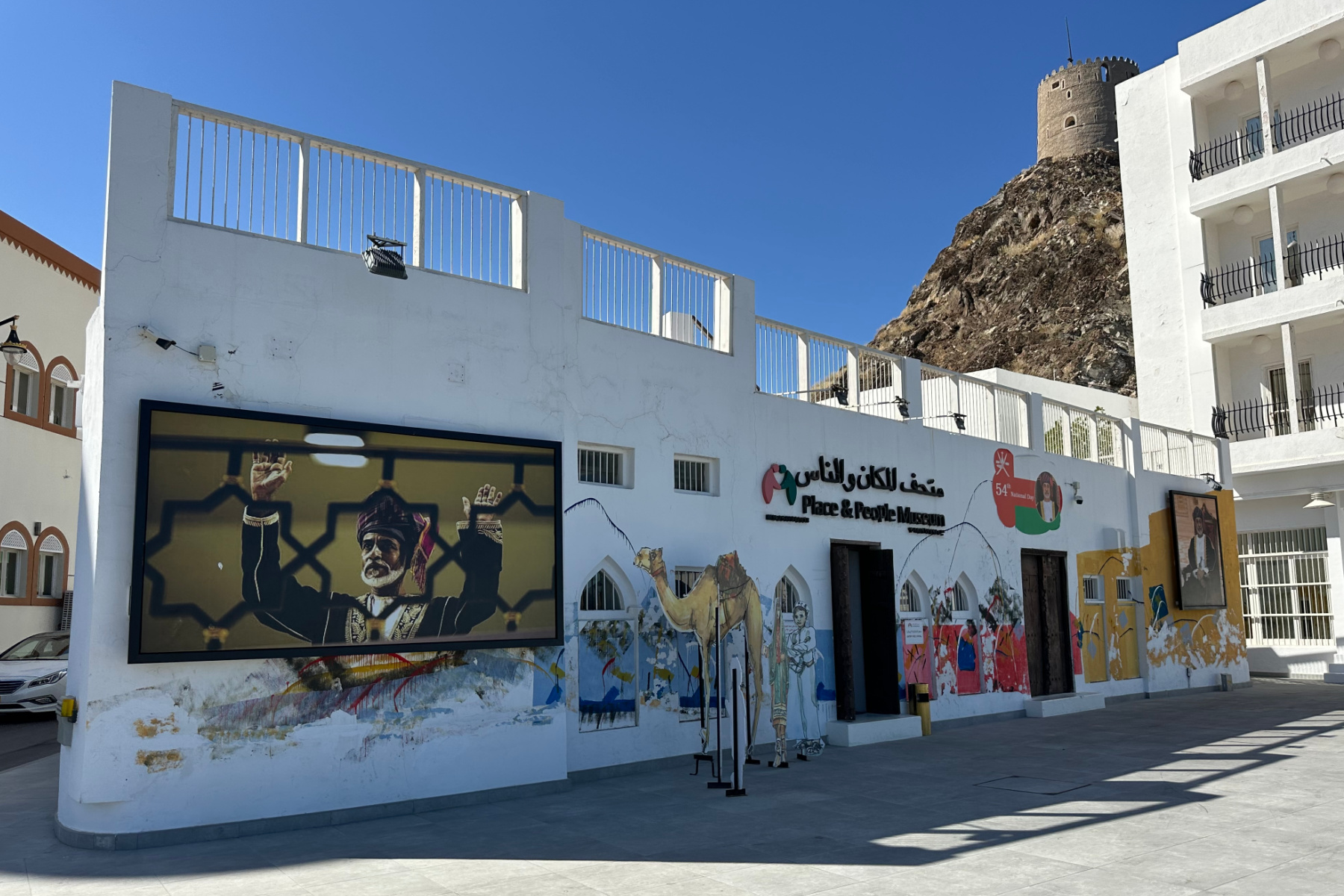 The outside of the Places and People Museum in Muttrah. It is a white building with pictures of Sultak Qaboos and Sultan Haitham bin Tariq Al Said on the front