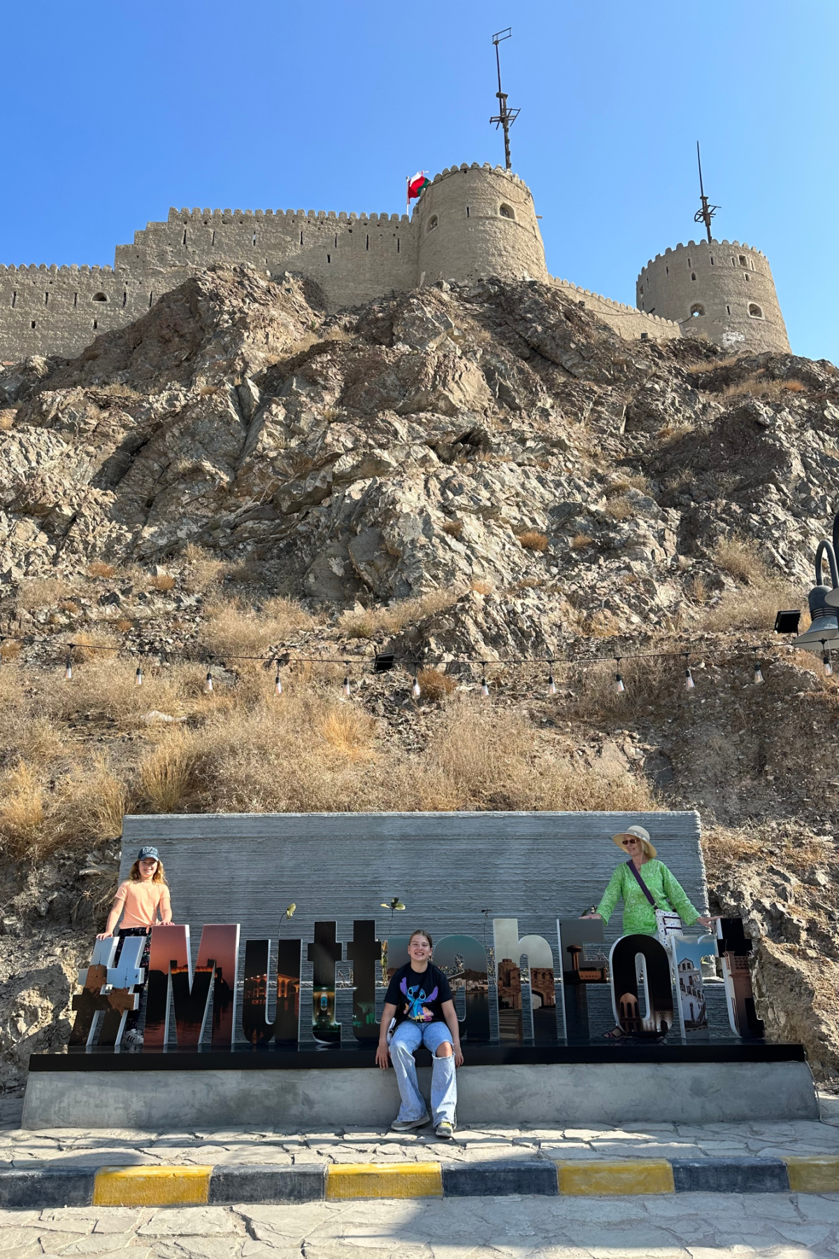 My children and mother stood with a large sign spelling out Muttrah Fort below the fort itself